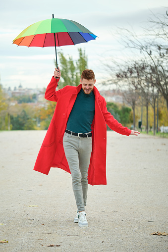 Latin young man with a red coat and rainbow umbrella walking on balance in the street in autumn.