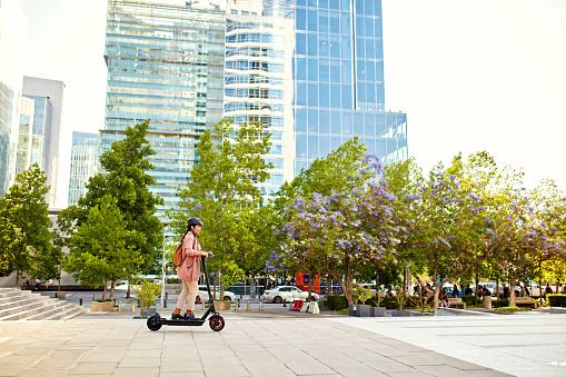 Full length side view of environmentally aware professional wearing safety helmet and riding electric push scooter through office park esplanade.