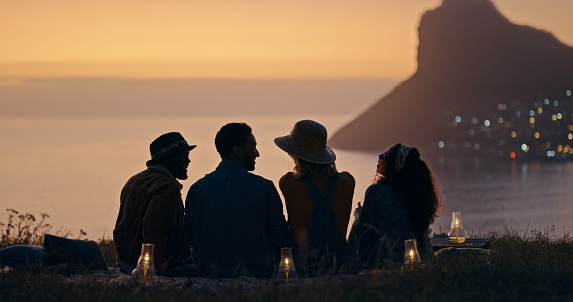 Group of people with beach view at night outdoor holiday, adventure and relax together on ground in silhouette rear. Social friends talking in shadow on mountains, nature and sunset with city bokeh