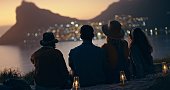 Beach view with group of people at night for outdoor travel, adventure break and relaxing on ground together in rear. Social friends silhouette on mountains or nature for sunset in city bokeh lights