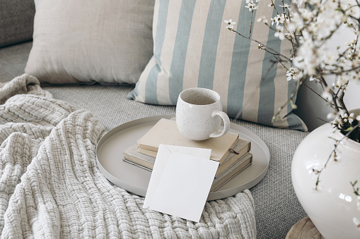 Spring breakfast scene. Blank greeting card mockup. Cup of coffee, tea on books. Round beige tray. Blossoming cherry plum tree branches in ceramic vase. Cozy linen sofa, cushions, Easter still life.