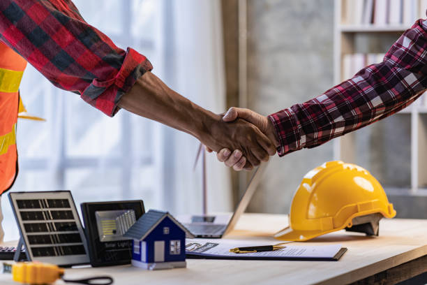 istock Yellow hard hat on table and house model solar windmill with construction team handshake to start new project contract plan in renewable energy concept contractor office center 1477109080