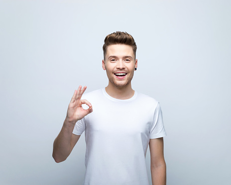 Happy young man wearing white t-shirt smiling at camera and showing ok sigh. Studio shot, grey background.