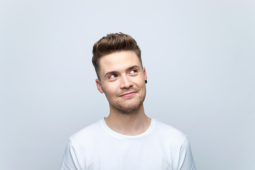 Headshot of pleased young man wearing white t-shirt looking up at copy space. Studio shot, grey background.