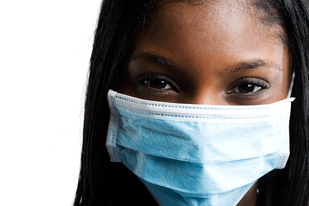 Twenty-something female doctor with blue surgical mask, gloves, scrubs and safety glasses, removing mask, isolated on white