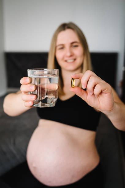 oméga 3. la femme enceinte prend des vitamines tous les jours et de l’eau en verre. les mains féminines tiennent des suppléments ou des vitamines. fille assise dans son lit à la maison. grossesse, traitement, médicaments concepts de soins de santé. - fatty acid photos et images de collection