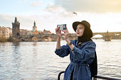 Stylish young woman traveller wearing black hat sitting taking pictures with her smartphone on Vltava river shore in Prague with Charles Bridge on background.