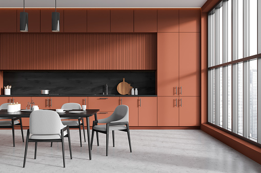 Stylish kitchen interior with dining table and chairs, grey concrete floor. Cooking corner with shelves and kitchenware, panoramic window on skyscrapers. 3D rendering