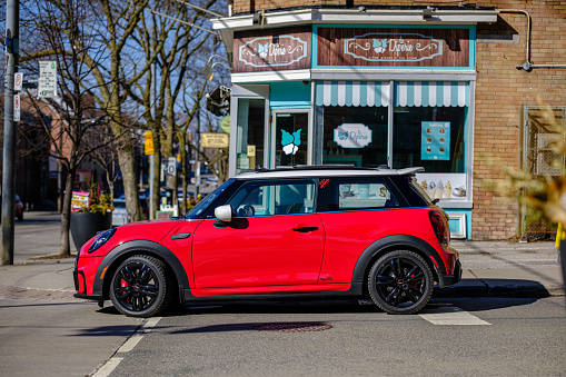 Toronto, Ontario, Canada- March 26, 2023. Chili red colour MINI COOPER on the spring sunny street  in Toronto East side, Canada. This is the third generation model F56 JCW, since BMW took over iconic brand of MINI. MINI featured in the photo is John Cooper Works model, the most powerful 2 door version. For the first time, this compact car features engine build and designed by BMW, and packs even more power and torque than previous models since 2002 to present. Original design clues and themes are still present on this brand new model. Mini has been around since 1959 and has been owned and issued by various car manufacturers.