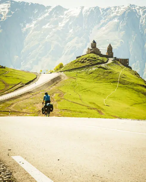 Male cyclist on touring bicycle cycling towards Gergeti trinity church with mountains background. Cycling holidays and travel around caucasus mountain range.