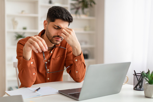 Tired young Middle Eastern man freelancer working from home office, using laptop, free copy space. Fatigued guy sitting at desk in front of pc computer, holding taking off glasses, rubbing nose bridge