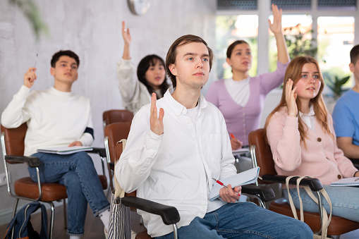 High school students sitting on chairs and raising hands to answer in lecture room