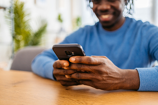 Close-up of a Smiling young black handsome man relaxing at home, sitting at the kitchen table and texting on smartphone.