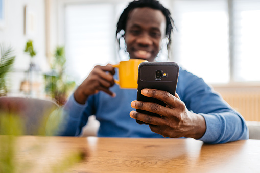 Smiling young black handsome man relaxing at home, sitting at the kitchen table, drinking coffee and texting on smartphone.