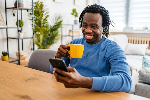 Smiling young black handsome man relaxing at home, sitting at the kitchen table, drinking coffee and texting on smartphone.