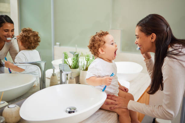 mom teaching baby to brush its teeth, on the bathroom counter in home and a clean smile on her face. healthy oral hygiene for kid means using child friendly toothpaste, toothbrush and dental routine - offspring child toothy smile beautiful imagens e fotografias de stock