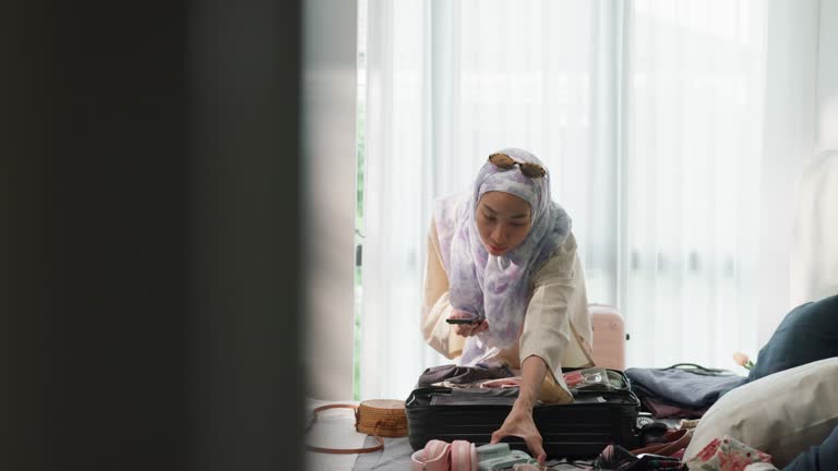 Young Asian muslim woman preparing suitcase use smartphone checking list accessories in bedroom at home. Travel vacation.