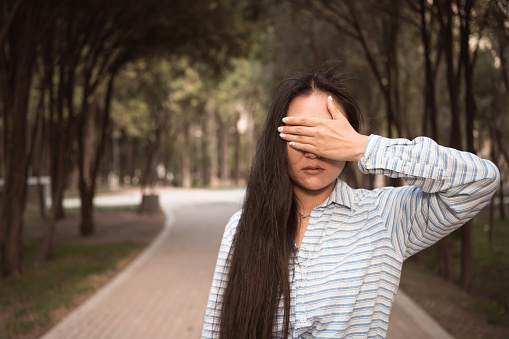 brunette woman covering by hand her eyes standing in city park