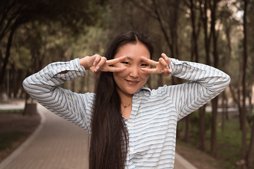 Asian young woman gesturing victory by hands and posing for photo outdoors