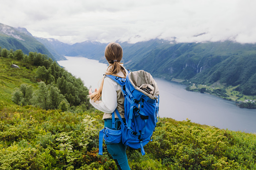Female backpacker with long hair, with cute pack in blue backpack contemplating a trip with view of the scenic fjord in Western Norway, Scandinavia
