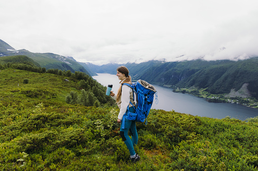 Female backpacker with long hair holding reusable cup, with cute pug in blue backpack contemplating a trip with view of the scenic fjord in Western Norway, Scandinavia