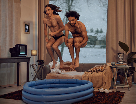 Hipster, rebel and jump into inflatable pool in a lounge inside a retro home or house having fun and playing. Funny, snow and men dive together in the living room for a winter vacation or holiday