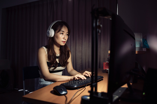 With her computer open in front of her, an Asian female student actively participates in an online evening course, wearing noise-canceling headphones to block out distractions and listening intently to the professor's lecture.