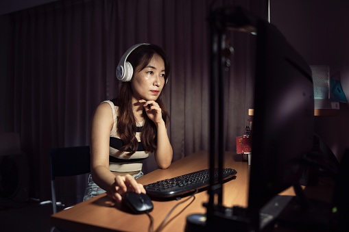 With her computer open in front of her, an Asian female student actively participates in an online evening course, wearing noise-canceling headphones to block out distractions and listening intently to the professor's lecture.