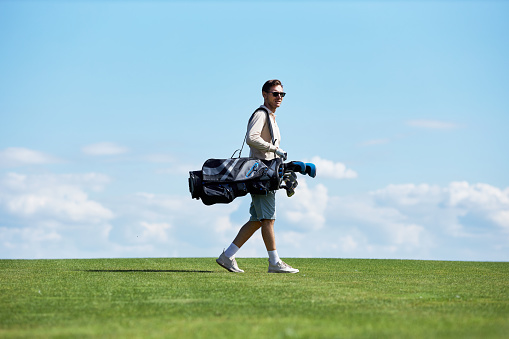 Minimal side view portrait of rich sporty man carrying golf bag walking on green field against sky, copy space