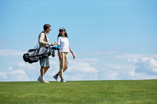 Minimal side view portrait of elegant sporty couple carrying golf bag walking on green field against blue sky, copy space