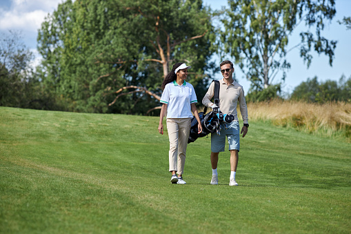Full length portrait of elegant sporty couple carrying golf bag walking on green field towards camera and chatting, copy space