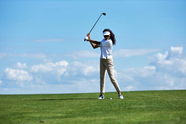 young woman playing golf on green field against sky and swinging golf club - golf swing golf golf club golf ball imagens e fotografias de stock