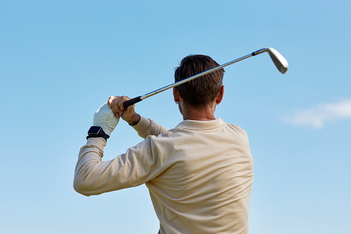 Back view of unrecognizable man swinging golf club against clear blue sky