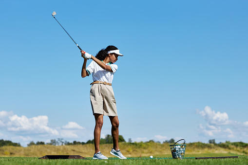 Side view portrait of sporty woman playing golf on green grass outdoors and swinging golf club, copy space