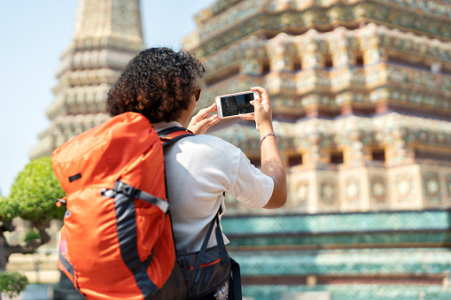 Temples are tourist attractions that calm the mind and feel relaxation. Back view of African America woman in causal cloth and orange backpack use smartphone take picture of temple. Tourist people travel alone and use phone record vlog on vacation trip in Thailand.