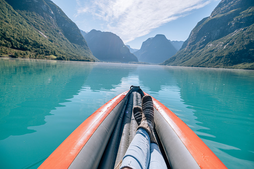 POv view of woman paddling on red inflatable canoe on peaceful environment in Norway. Blue lake and green mountains