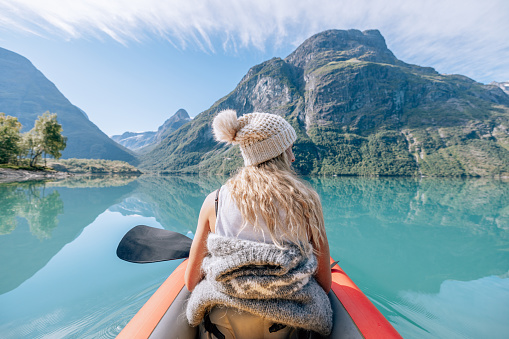 Rear view of woman paddling on red inflatable canoe on peaceful environment in Norway. Blue lake and green mountains