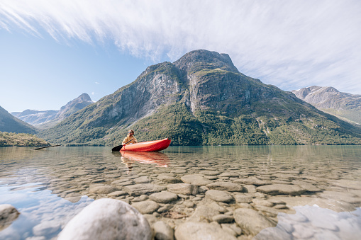 Active man paddling on red inflatable canoe on peaceful environment in Norway. Blue lake and green mountains