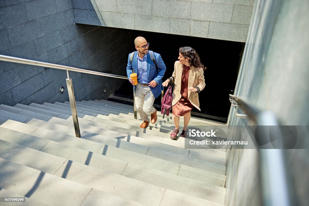 Smiling business partners walking up staircase High angle view of male and female colleagues in their 30s talking as they return to office park from appointment. Staircase Stock Photo