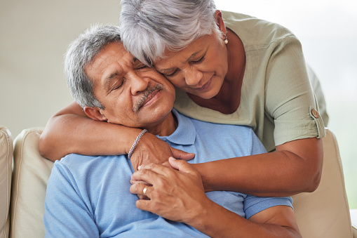Senior couple hug, comfort or console in a living room at home. Elderly husband and wife holding each other on sofa and embracing in lounge. Retired man and woman bonding and feeling love together