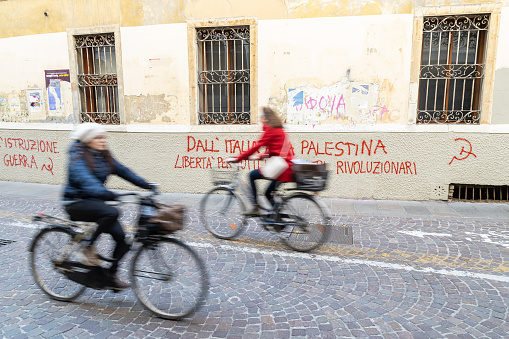 Padua, Veneto, Italy - Mar 11th, 2023: Blurred cyclists riding past left wing Pro-Palestine street art graffiti painted on the wall in Padua city center