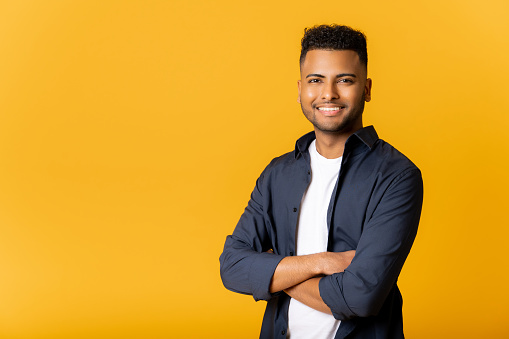 Confident young indian man standing in confident pose with arms crossed, looking at camera, copy space for advertising new promotion, arab guy standing isolated on yellow background.