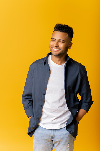 Portrait of carefree indian guy in casual shirt standing isolated on yellow background, arab man posing in studio, holding hands in pockets of jeans, relaxed serene handsome man looking away