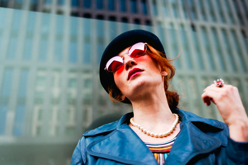 Portrait of a beautiful young woman in the city in front of a skyscraper. She is wearing red sunglasses.