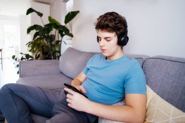 istock Caucasian teenage boy with acne problems in wireless headphones reading, messaging, using social media, playing games online using his smartphone sitting on the sofa. Online activity. Selective focus. 1477206458