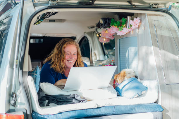 istock Smiling middle-aged woman working on laptop while lying in camper car vehicle with cockapoo puppy pet during road trip. Remote worker people. Enjoying free lifestyle, travel, vacation, freedom. 1477206514