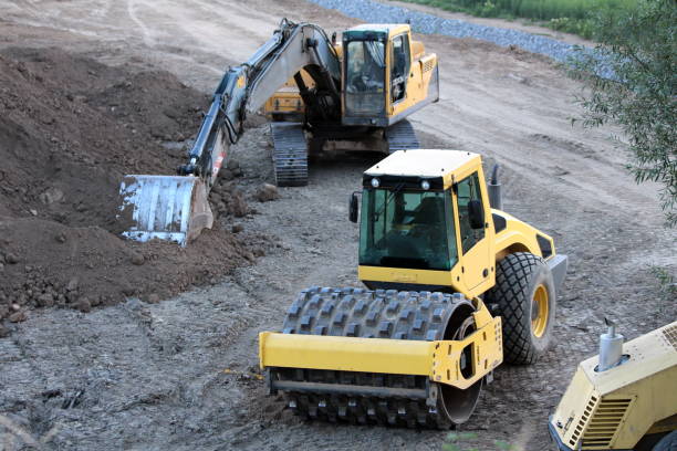 Industrial single drum roller used for medium to heavy soil compaction next to industrial crawler excavator at local road construction site surrounded with gravel and sand mixed with dirt on warm summer day at sunset