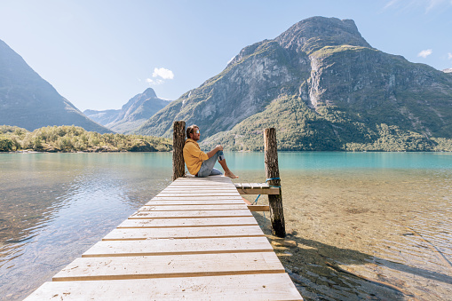 Man with red inflatable canoe on peaceful environment in Norway. Blue lake and green mountains.
He relaxes on a wooden pier.