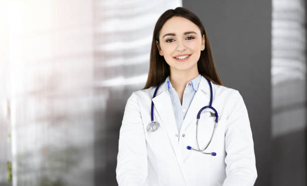 une jeune femme-médecin souriante se tient debout dans une clinique ensoleillée à l’intérieur - portrait doctor paramedic professional occupation photos et images de collection