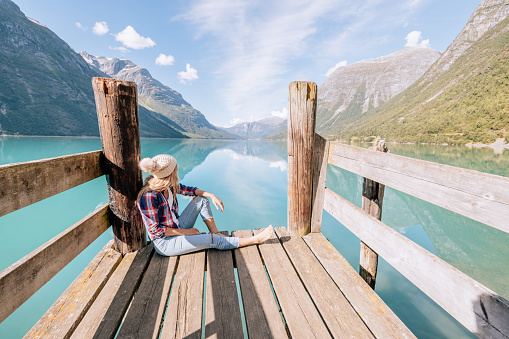 Woman with red inflatable canoe on peaceful environment in Norway. Blue lake and green mountains.\nShe relaxes on a wooden pier.